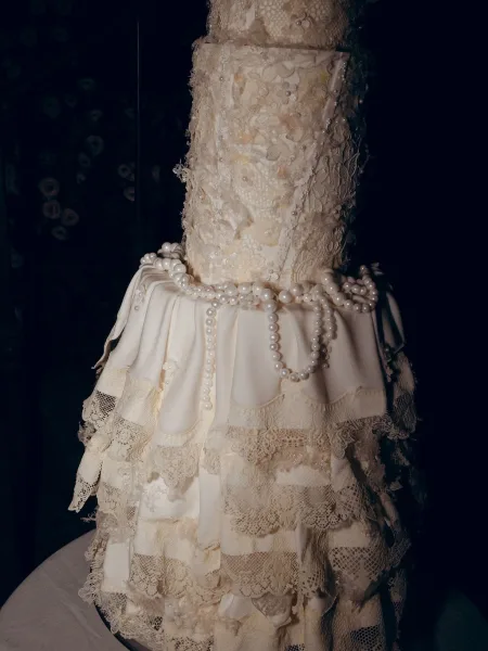 Wedding cake with lace wedding cake details, pearl garland, and fondant floral applique on a cake table against a dark backdrop