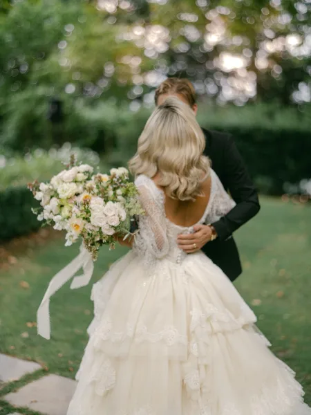 Couple portrait of bride and groom embracing during wedding first look, bride holding rose bouquet with ribbon streamers in garden bokeh light