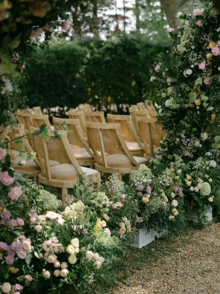 Ceremony aisle decor with a floral ceremony arch of roses and greenery, ground arrangements and wooden chairs along a gravel garden path