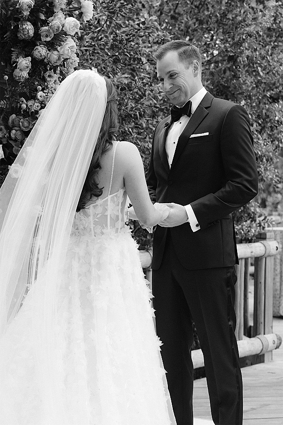 First look moment as bride in wedding dress and veil holds hands with groom in black tuxedo and bow tie by garden greenery railing
