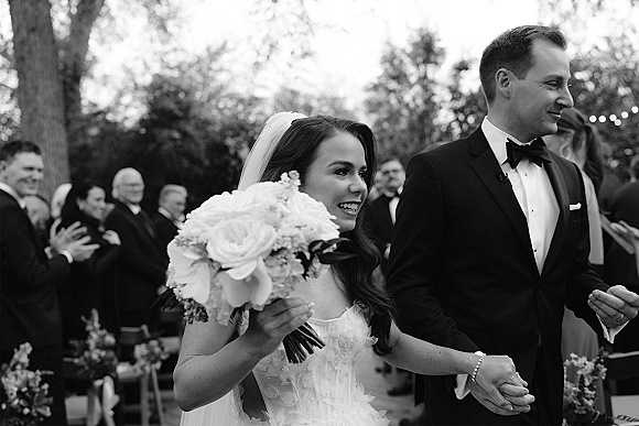 Recessional moment as bride and groom walk the outdoor aisle holding hands, bouquet and veil flowing, guests cheering under string lights
