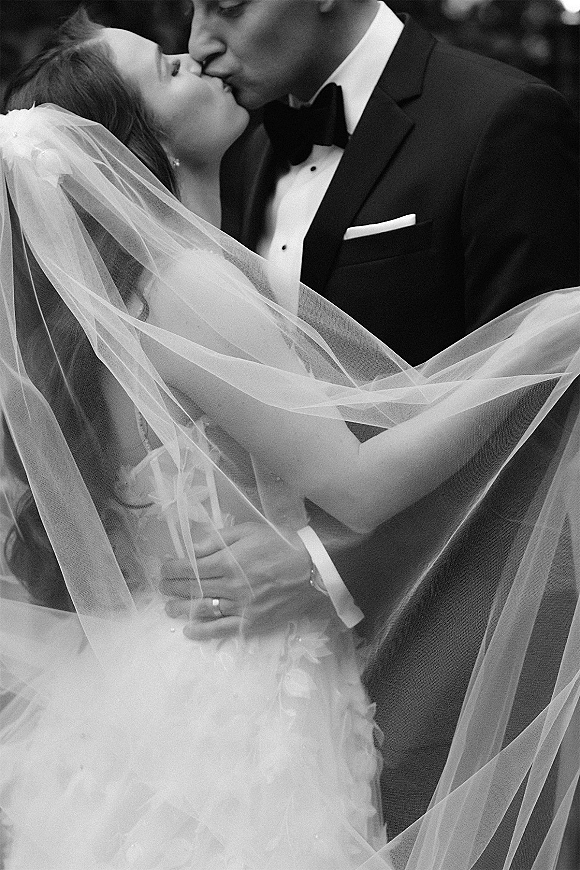 Wedding kiss portrait in black and white of bride and groom kissing under a veil, groom in tuxedo with bow tie, blurred crowd behind