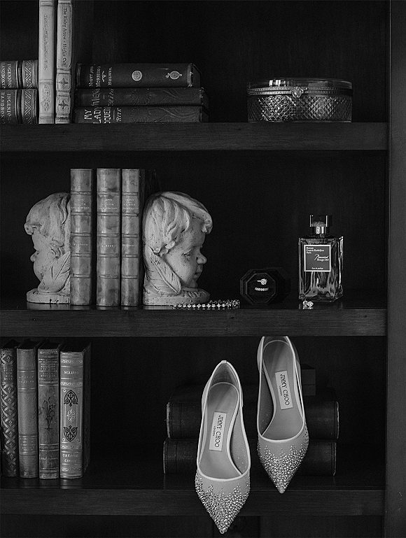 Bridal shoes in a wedding shoe flatlay with rhinestone accents, perfume bottle, bracelet, and jewelry box on stacked books by a bookshelf