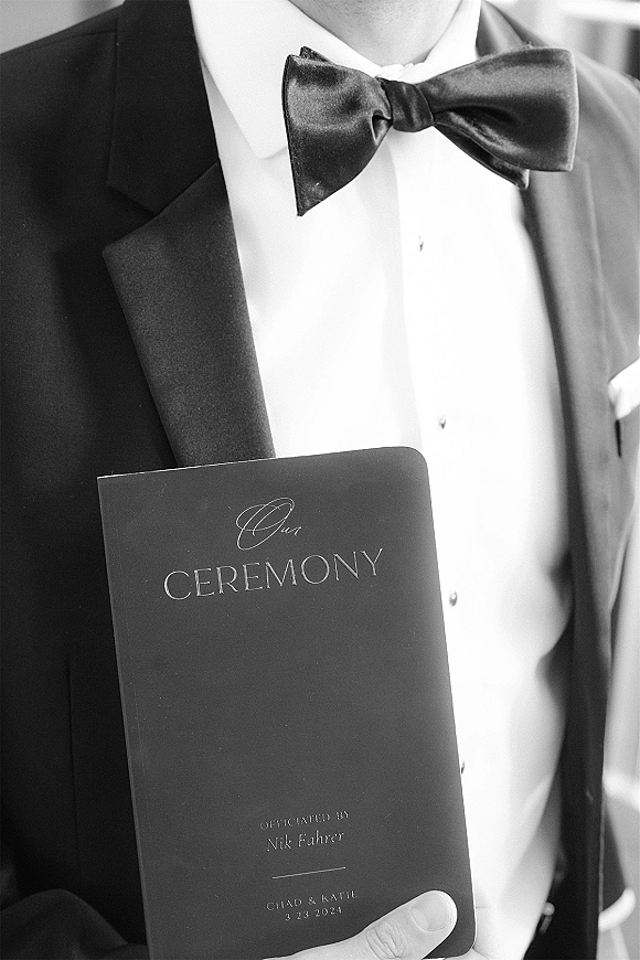 Wedding ceremony program held against a tuxedo jacket with satin bow tie, showcasing a ceremony program booklet in a neutral indoor setting