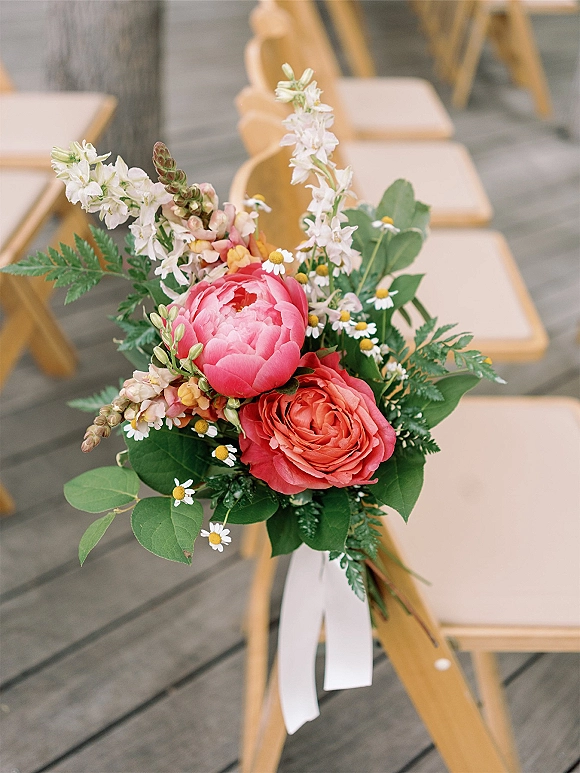 Wedding aisle bouquet with peonies and garden roses tied to a ceremony chair, white ribbon and greenery accents on a wood deck seating area