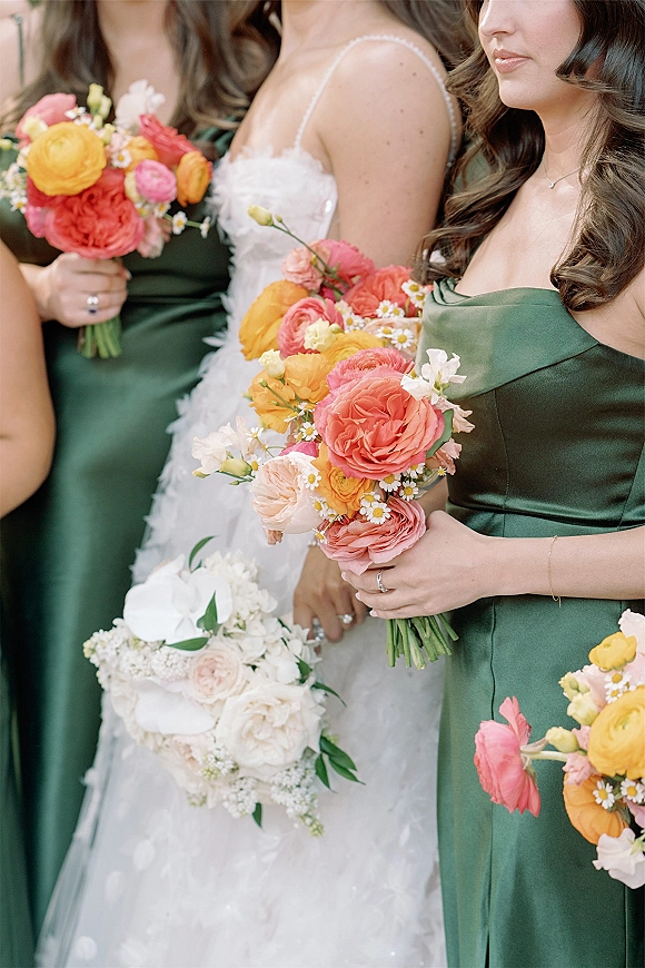 Bridesmaid bouquets in vibrant mixed flowers held beside a white bridal bouquet, paired with green satin dresses against a neutral indoor backdrop