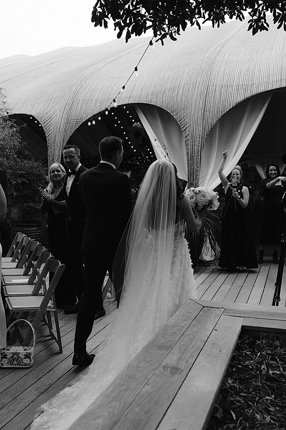 Wedding recessional with bride and groom walking away, her cathedral veil and bouquet trailing on a wooden deck under string lights as guests cheer