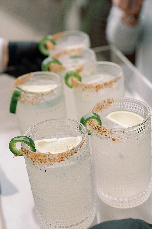Signature wedding cocktails with chili-salt rims and lime wedges on a tray, jalapeño slices floating in glasses on an outdoor patio table