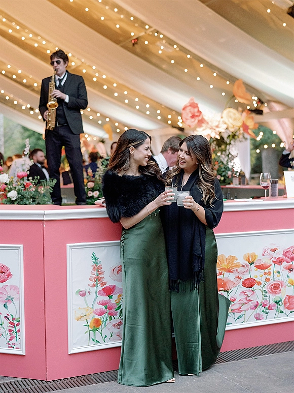 Wedding guest portrait of bridesmaids at bar holding cocktails in satin dresses, with pink floral bar panels and string lights in a tent