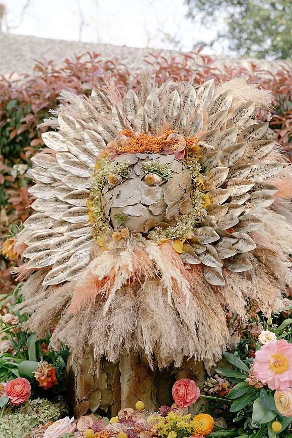 Escort card display with calligraphy escort cards on feather-shaped cards, framed by pampas grass, orchids, and ranunculus against a stone wall outdoors