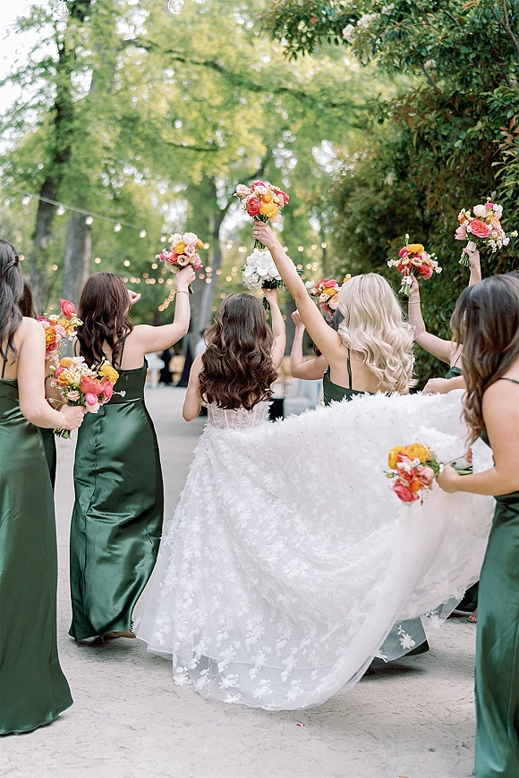 Bride with bridesmaids walking away as bridesmaids cheering photo, lifting colorful bouquets on a tree-lined garden walkway under string lights