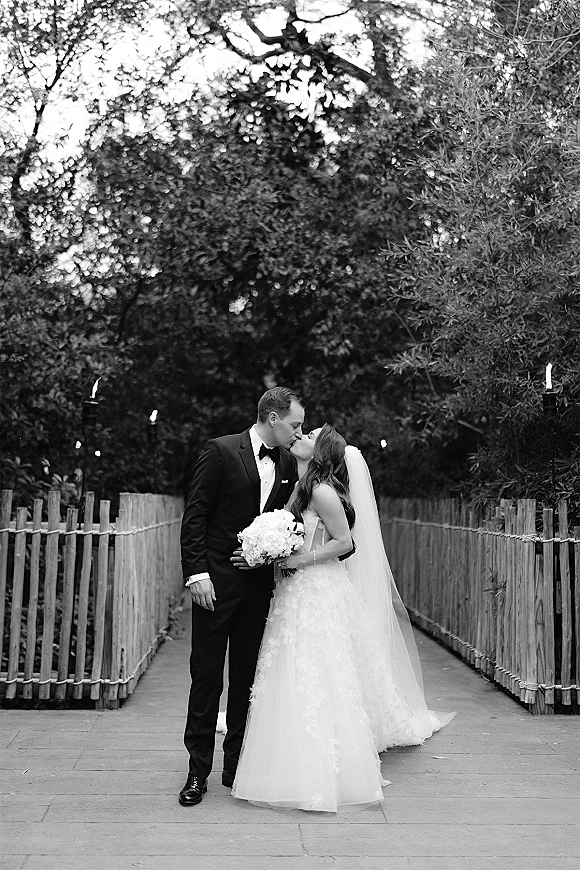 Wedding kiss in a black and white wedding photo, bride in strapless dress and long veil kissing tuxedo groom on a walkway with trees and fence