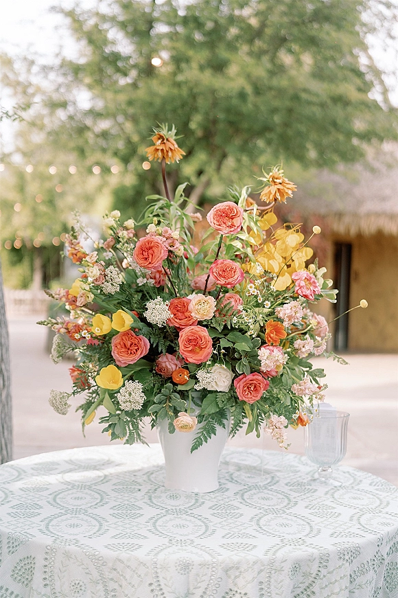 Wedding floral centerpiece in a white ceramic vase, bursting with orange and pink blooms on a table set under string lights outdoors