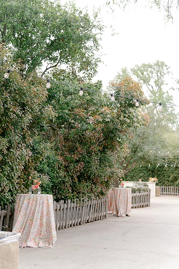 Cocktail hour decor with outdoor cocktail hour high-top tables, floral centerpieces and string lights along a garden walkway by trees and fence
