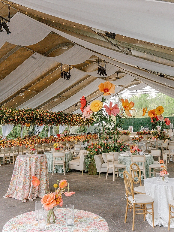 Reception decor with draped tent ceiling, string lights, hanging floral installations over round tables with candles and patterned linens inside tent