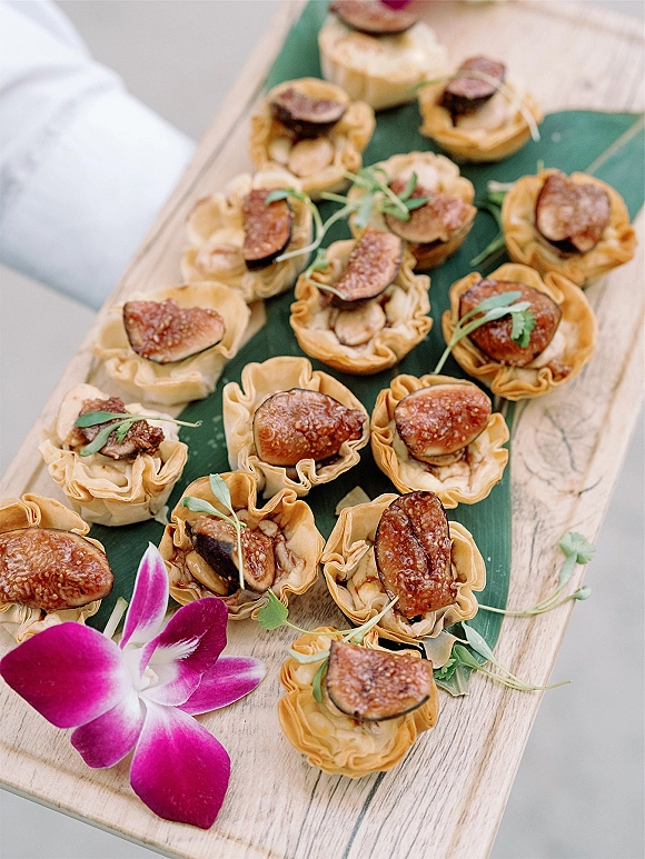 Wedding appetizers arranged as bite-size tartlets with fig slices and microgreens on a wooden serving board with an orchid accent
