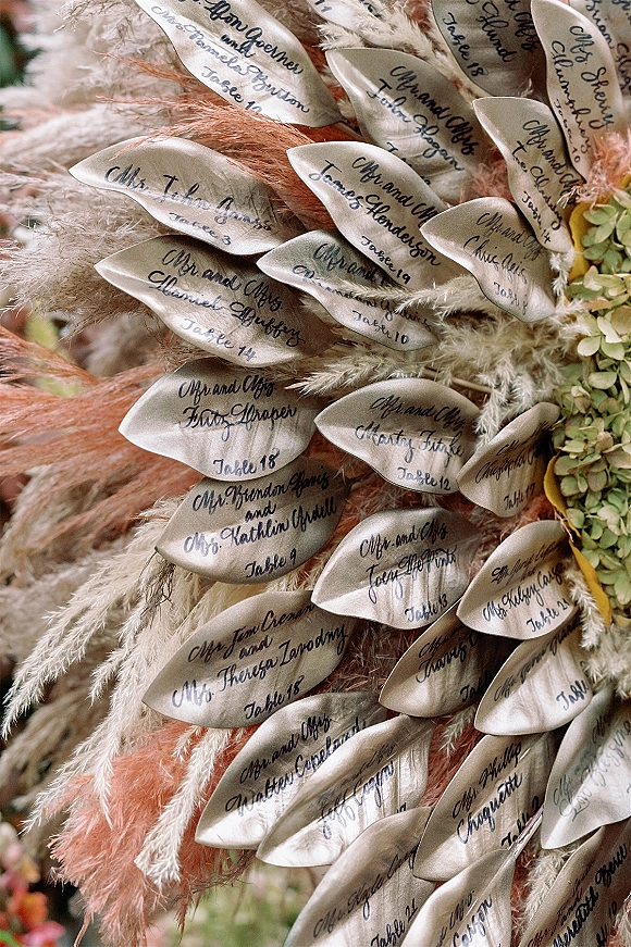 Escort card display with wedding escort cards in handwritten calligraphy on oval cards, accented by pampas grass and green hydrangea against blurred greenery