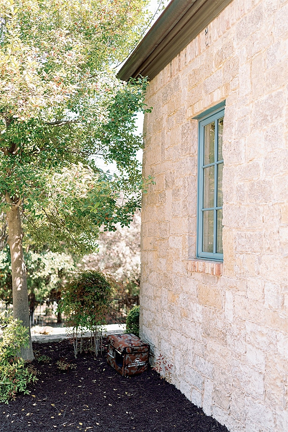 Stone house exterior with limestone wall exterior, blue-framed window and brick sill, bordered by mulch, shrubs, and a small tree in sunlit garden corner