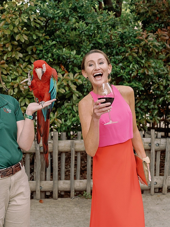 Wedding guest portrait of a woman in a colorblock wedding guest dress, laughing with a wine glass by green shrubbery and a fence