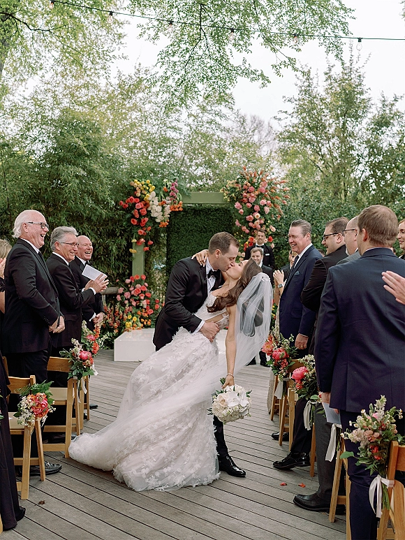 Ceremony kiss moment as bride in flowing veil dips with groom in tuxedo, bouquet in hand, under floral arch on a garden deck aisle
