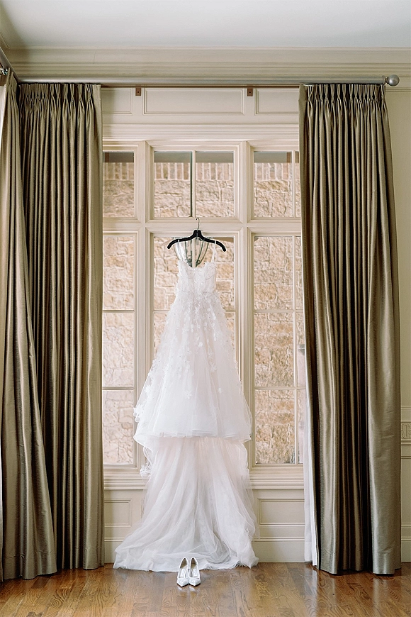 Wedding dress hanging by a large window, lace appliques and tulle skirt draping above white bridal heels on a hardwood floor