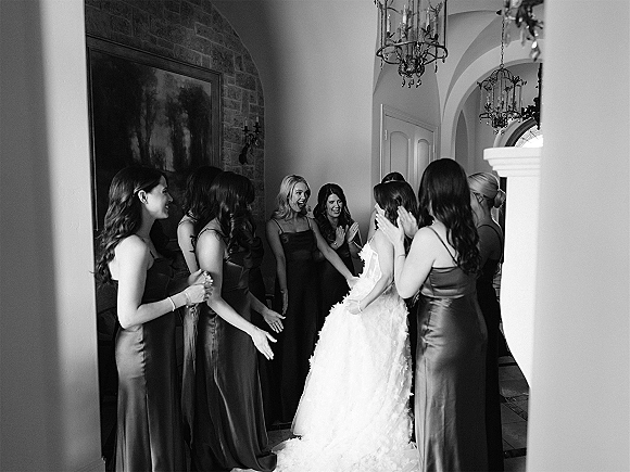 Bridesmaid reveal as women in dark satin dresses clap and smile at the bride in a textured gown in an arched hallway with chandeliers