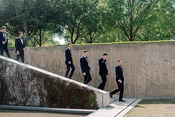 Groomsmen portrait of a group in black tuxedos and bow ties, walking in formation beside a concrete wall on an outdoor ramp with greenery