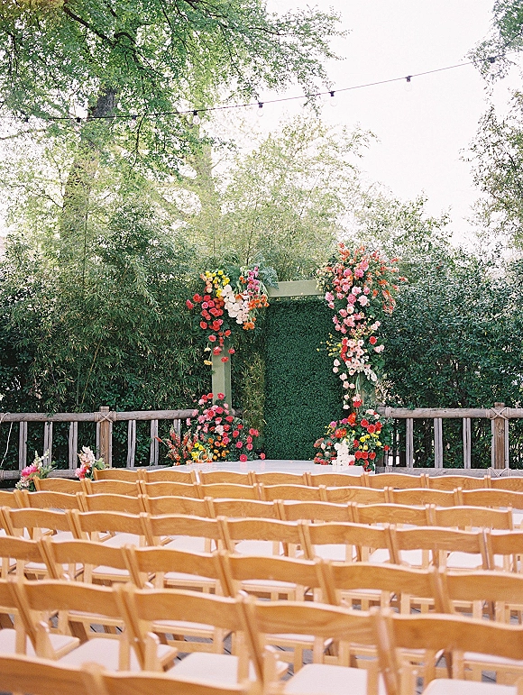 Ceremony setup with wood folding chairs facing a floral arch on a greenery wall backdrop, accented by string lights in a garden space
