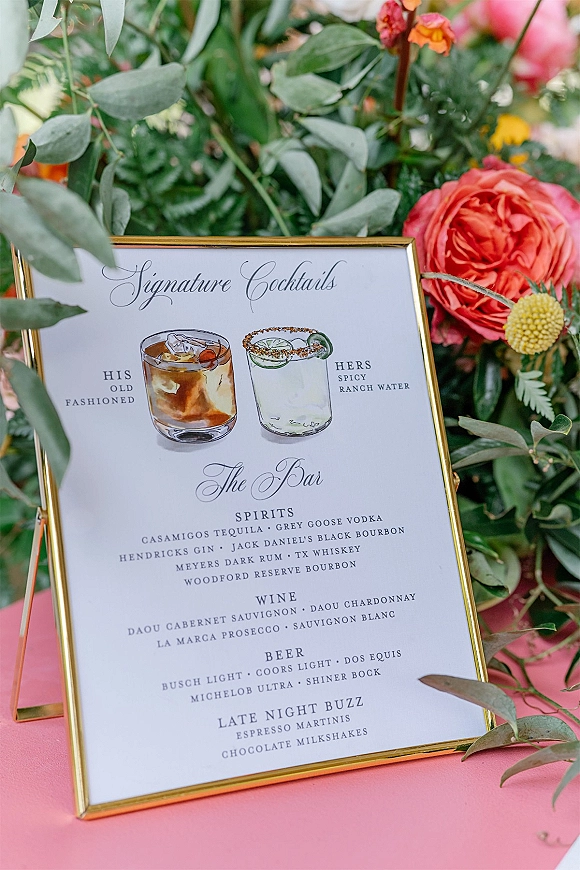 Signature cocktail sign in a gold frame with illustrated drink drawings, bar menu card, and rose greenery accents on a pink tabletop