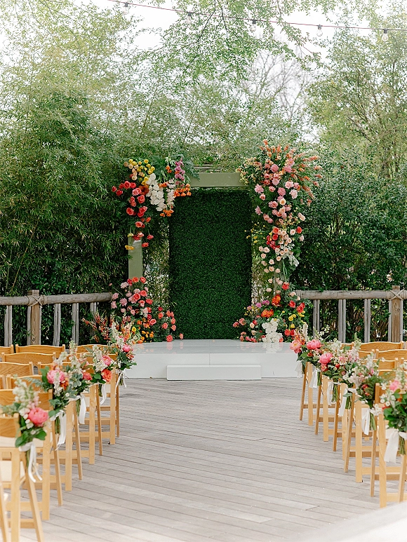 Ceremony setup with an asymmetrical floral arch and greenery wall backdrop, wooden chairs with ribbon streamers on an outdoor deck in garden trees