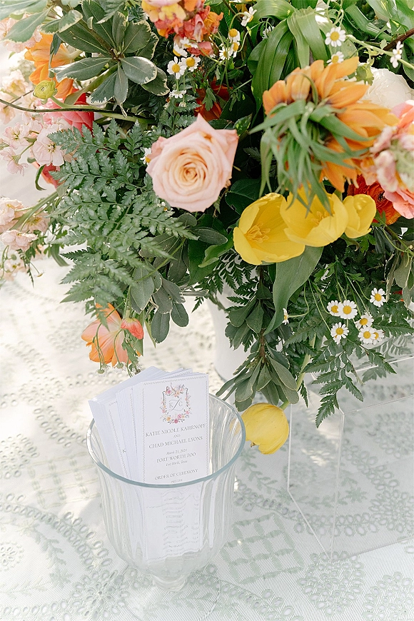 Wedding floral centerpiece of roses, tulips, and daisies with greenery on a clear acrylic stand beside wedding programs on lace tablecloth
