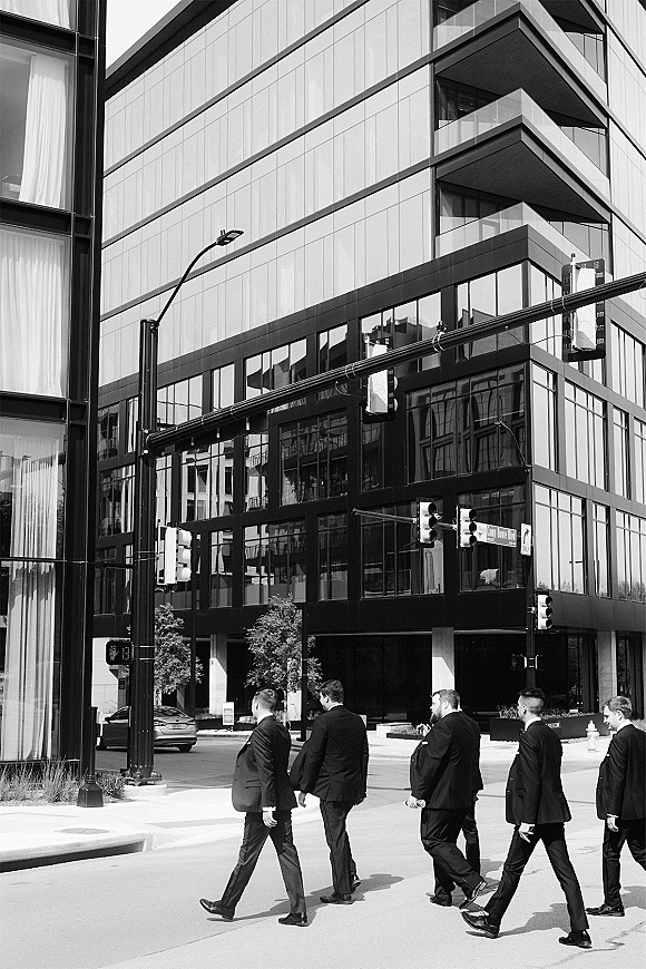 Groomsmen photo of men in black suits walking across a city crosswalk, modern office building with glass windows and traffic lights behind them