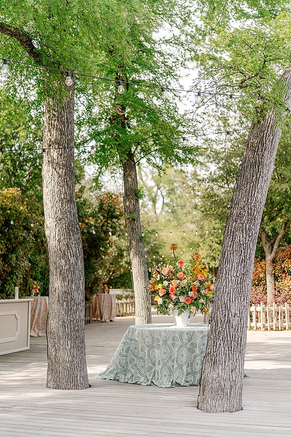 Outdoor wedding decor with a wedding sweetheart table featuring a white vase floral centerpiece on a lace tablecloth under string lights on a wooden deck