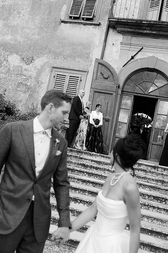 Wedding candid moment of bride and groom holding hands, looking back on stone steps by an arched doorway as guests watch behind them