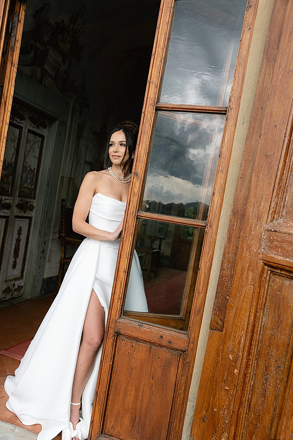 Bridal portrait of a bride in a strapless wedding dress with thigh slit, pearl necklace, and updo, standing by a wooden door with window panes