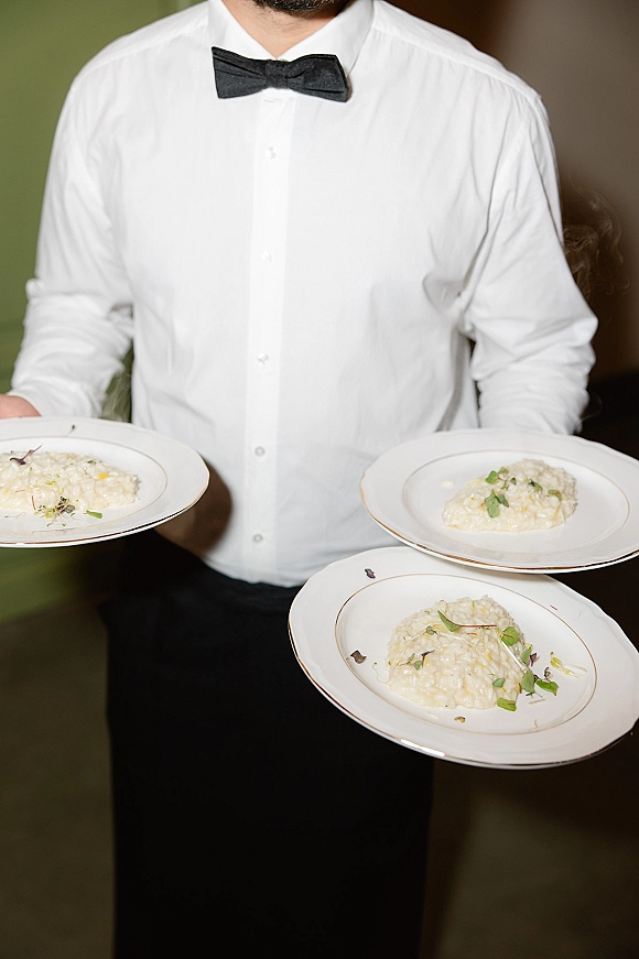 Wedding catering service waiter in black bow tie carrying plated wedding dinner of risotto with microgreens through an indoor venue doorway