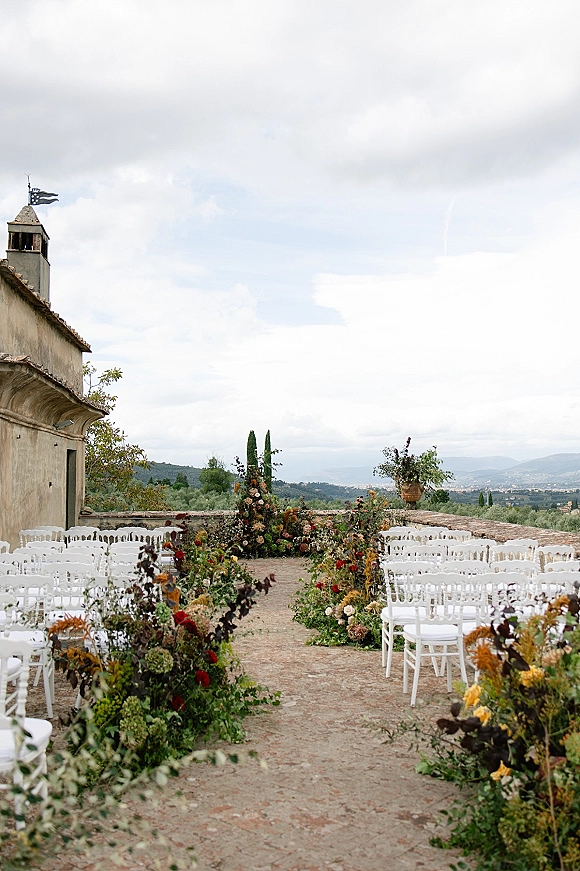 Ceremony aisle decor with white chairs and floral-lined aisle arrangements on a stone terrace facing a historic facade and misty mountains