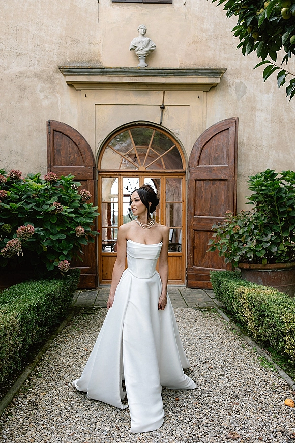 Bridal portrait of a bride in a strapless wedding dress with a layered pearl necklace and updo, standing by an arched doorway in a courtyard garden