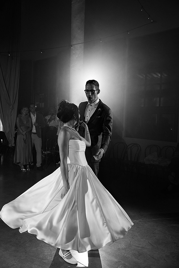 First dance as bride twirls her strapless gown, groom in glasses watches under string lights in a softly backlit reception hall