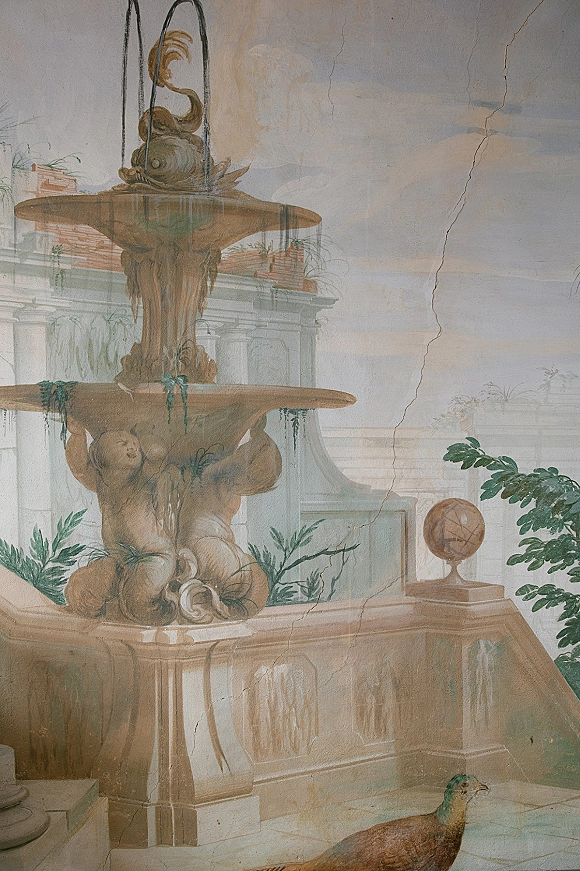 Wedding venue mural showing a hand painted wall mural of cherubs at a fountain, with a peacock motif on cracked plaster courtyard wall
