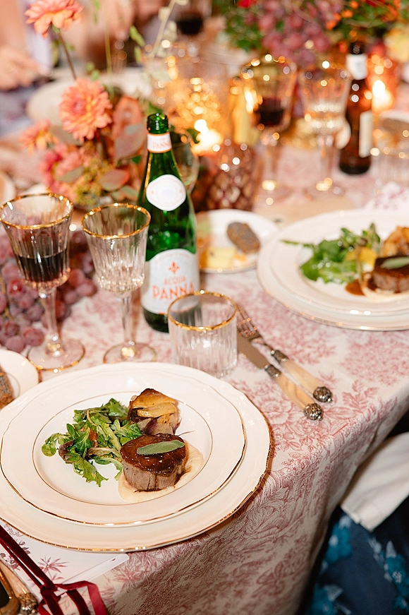 Reception tablescape with wedding place setting, pink toile tablecloth, gold-rim glassware, taper candles, floral centerpiece, blurred guests behind
