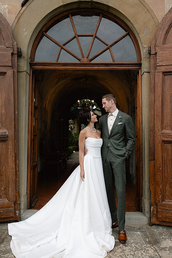 Couple portrait of bride and groom holding at the waist, gazing at each other in a stone arch doorway with wooden doors and chandelier