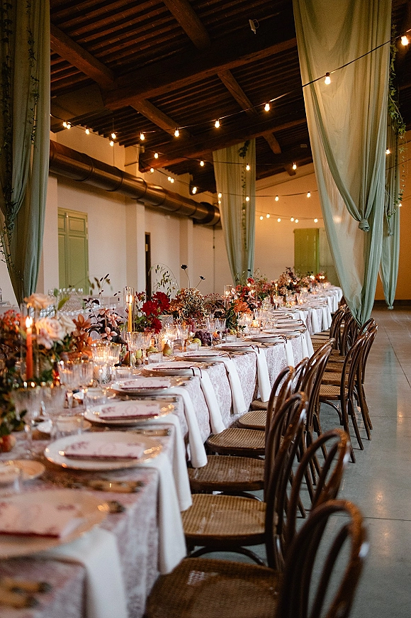 Reception tablescape on a long banquet table wedding setup with floral centerpieces, taper candles, fruit, and string lights in a wood-beam hall