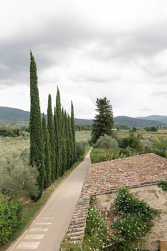 Countryside landscape with a cypress tree lined road leading past a stone farmhouse with flowering vines, set among rolling hills under clouds