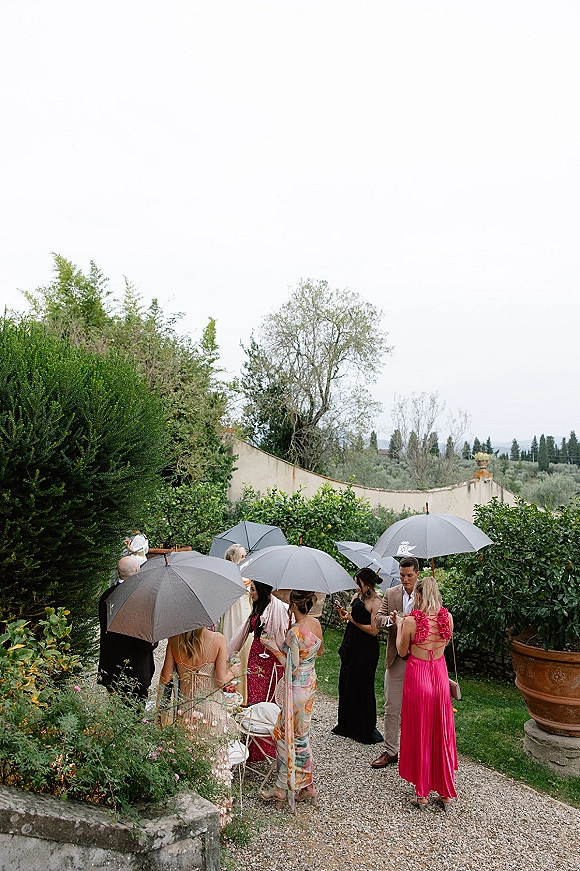 Wedding guests with umbrellas mingle in dresses and suits, holding cocktail drinks along a gravel garden path under an overcast sky