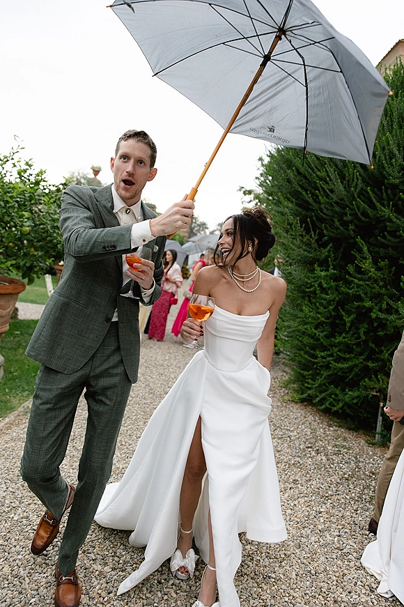 Wedding couple portrait of bride and groom under an umbrella, smiling with cocktails on a garden gravel path as guests watch behind