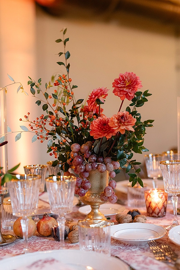 Reception tablescape with wedding table centerpiece in a gold compote vase, grapes, pomegranates, candles, and crystal glasses on patterned cloth indoors