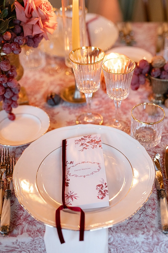 Wedding tablescape with wedding place setting, white plates topped with a menu card and burgundy ribbon, crystal glasses, and taper candles