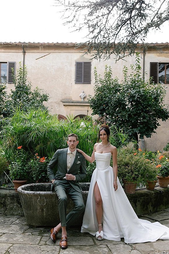 Couple portrait of bride in a strapless slit gown with long train, hand on groom’s shoulder in a garden courtyard with potted plants