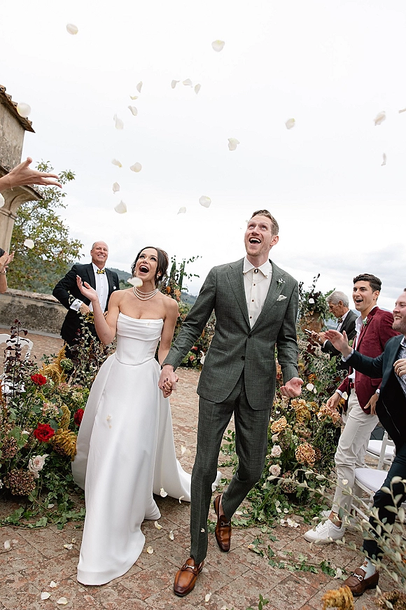 Wedding recessional with a rose petal toss as the bride and groom hold hands on a brick walkway in an outdoor courtyard with stone building behind